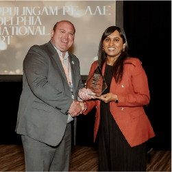 Man in grey suit smiling on left, shaking a smiling woman's hand who is wearing a red jacket and holding an award